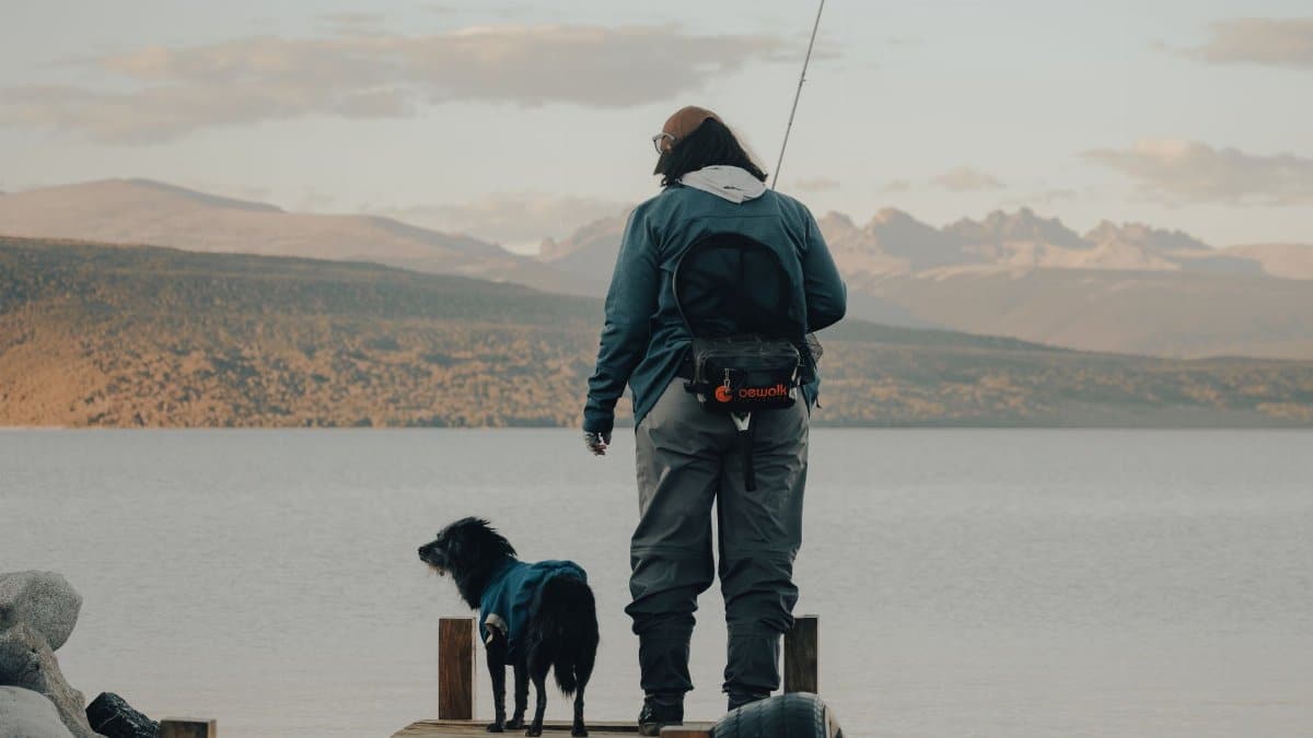 A man stands fishing with his dog on a tranquil boardwalk by a lake, surrounded by mountains.