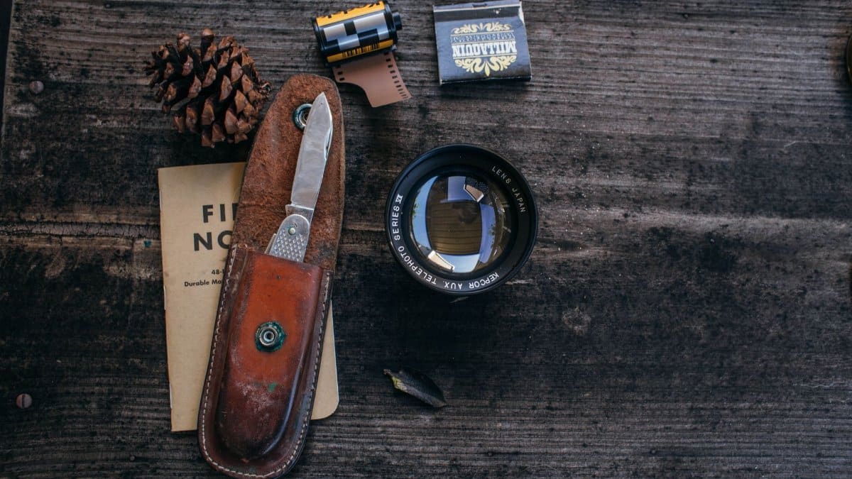Overhead of knife near bump and films near lens near small notepad and matchbox on wooden surface
