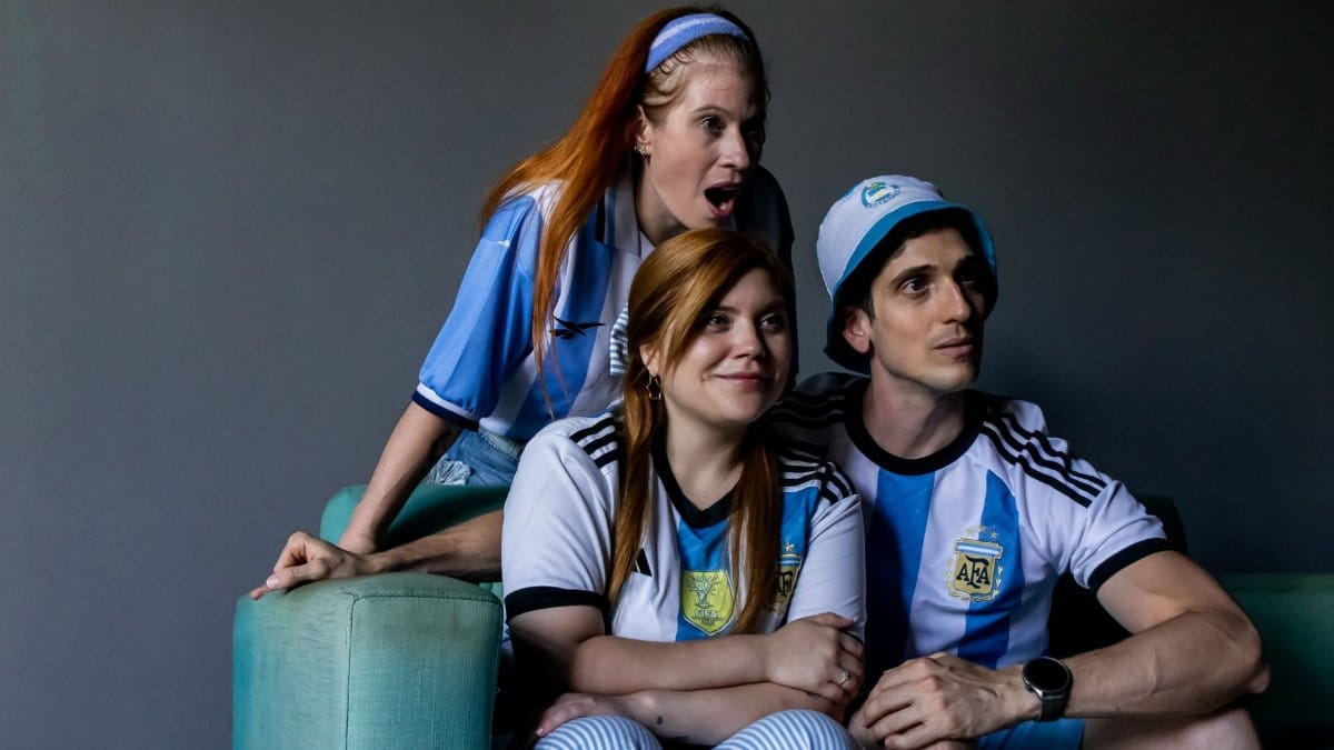 Three fans in Argentina jerseys watch a soccer match with excitement indoors.