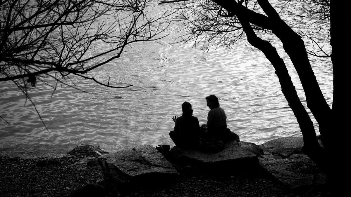 Two people sit on rocks under trees beside a calm river in Basel, Switzerland, creating a serene and reflective scene.