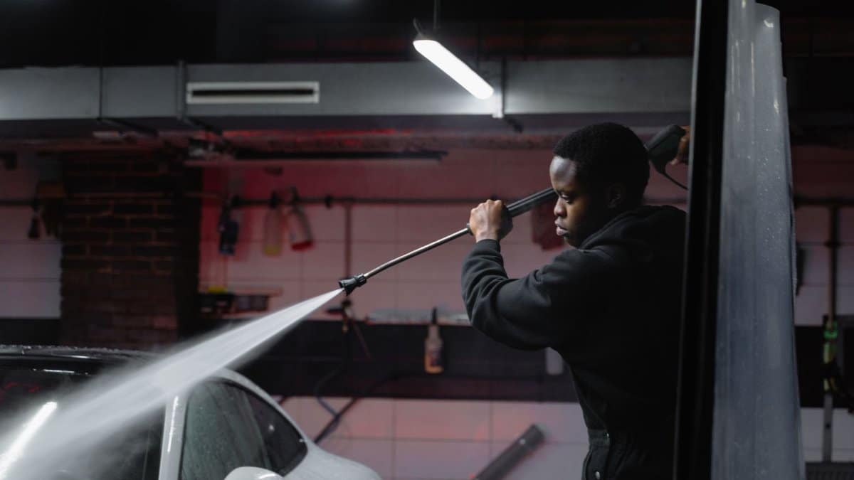 A man uses a high-pressure hose to wash a car in an indoor garage setting.