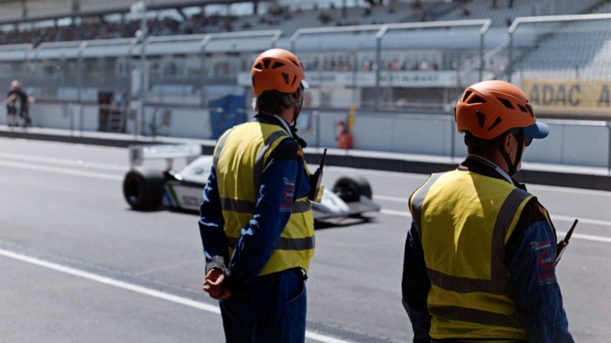 Two race officials in protective gear watch a car on the racetrack at a racing event.