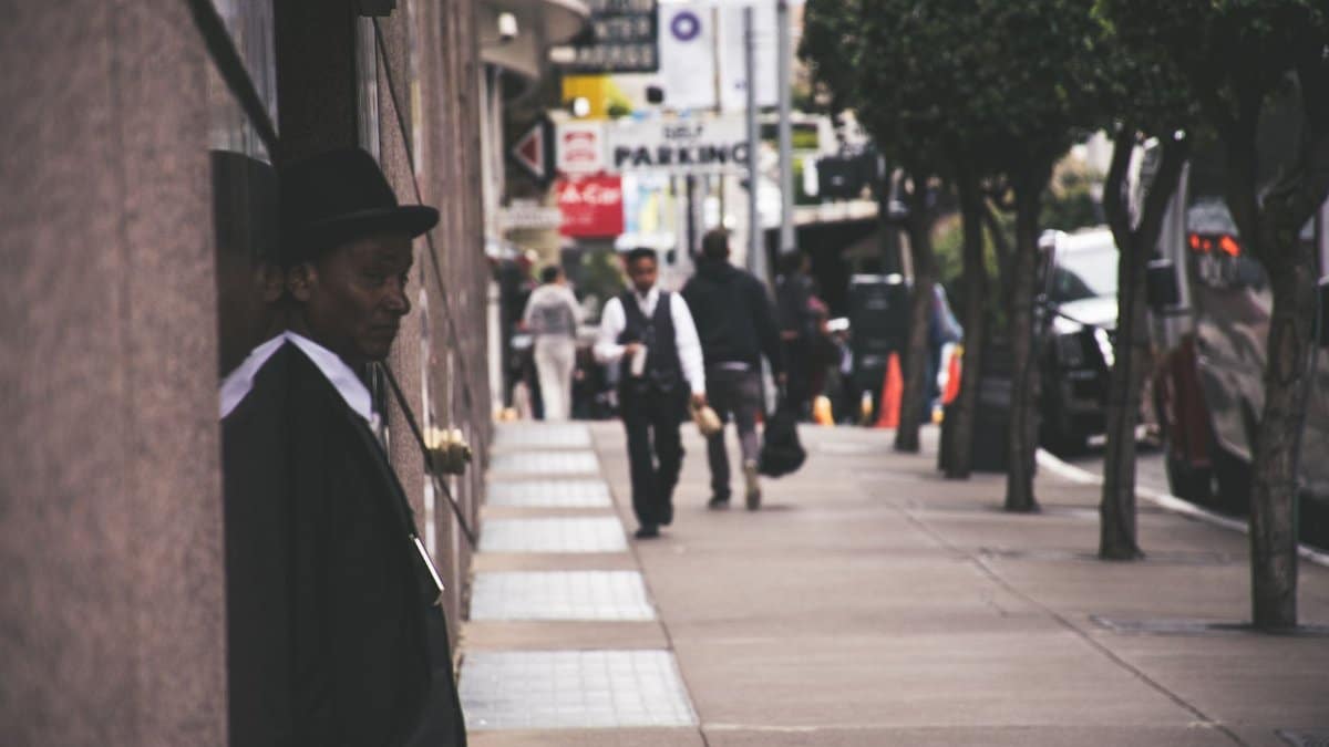 A man in a suit and hat leans against a city building, observing pedestrians.