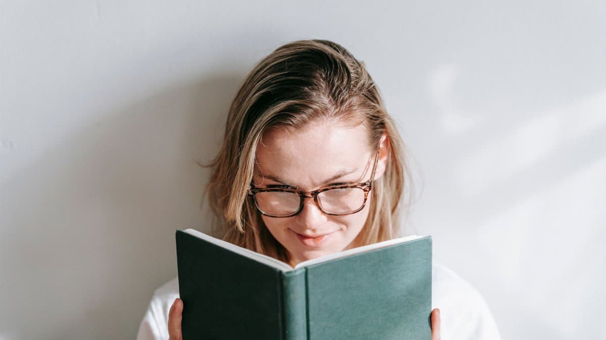 Interested young woman in glasses reading textbook while studying in sunshine on light background