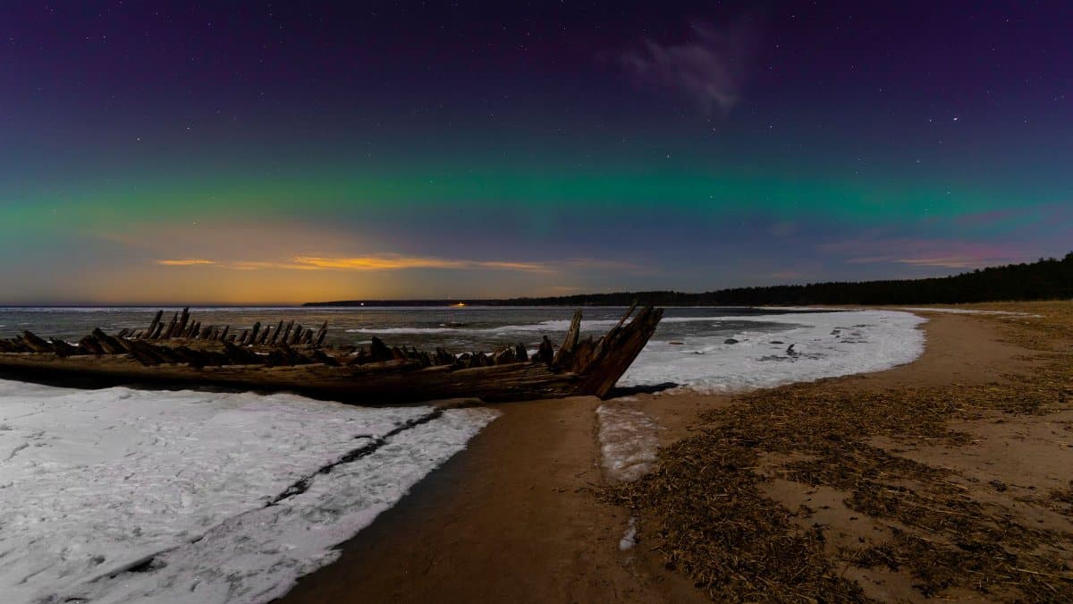 Beautiful northern lights arch over a frozen beach with shipwreck remains.