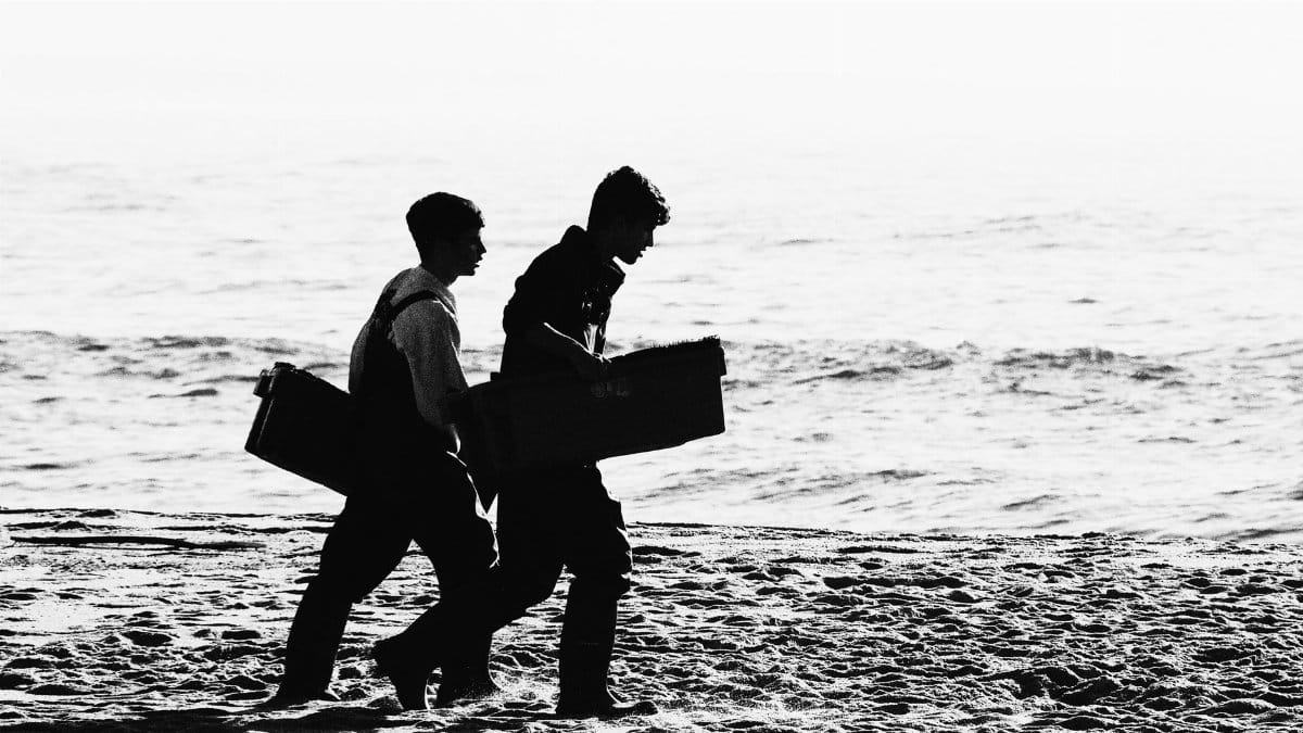 Black and white image of two adults carrying boxes along a beach shore.