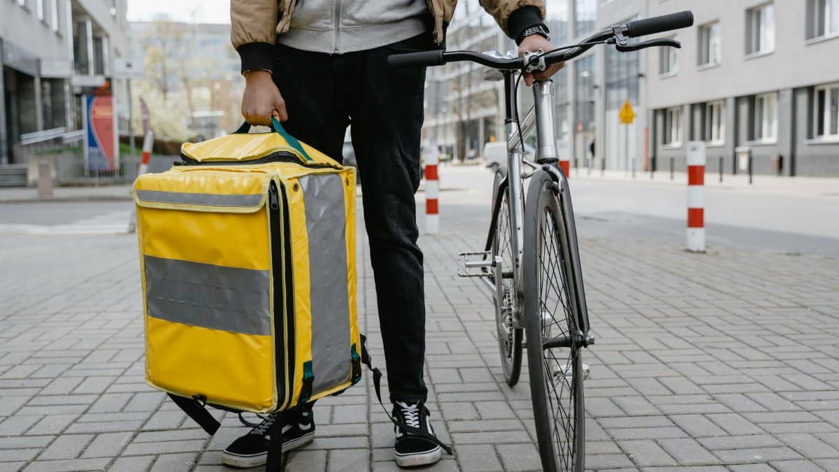 A cyclist with a yellow thermal bag stands with his bike on a city street, ready for food delivery.