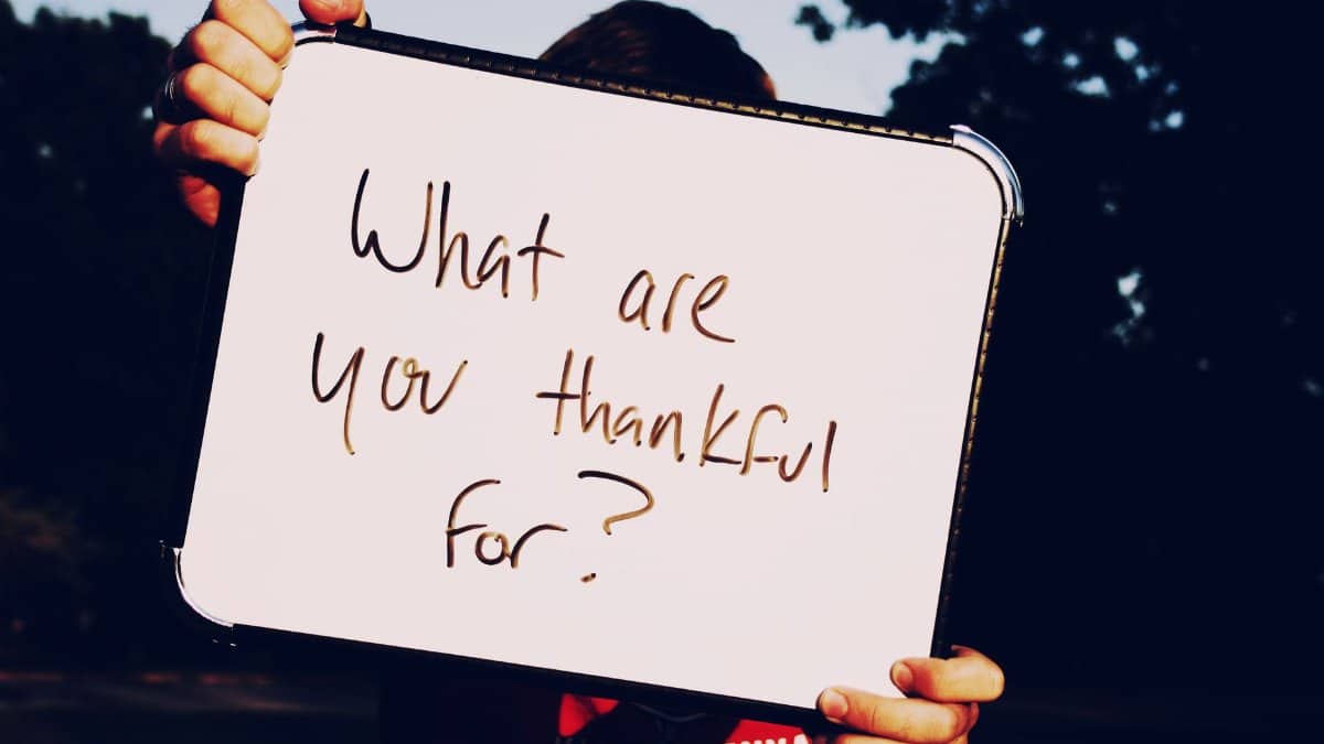 A person holds a whiteboard asking 'What are you thankful for?' against a dark outdoor background.