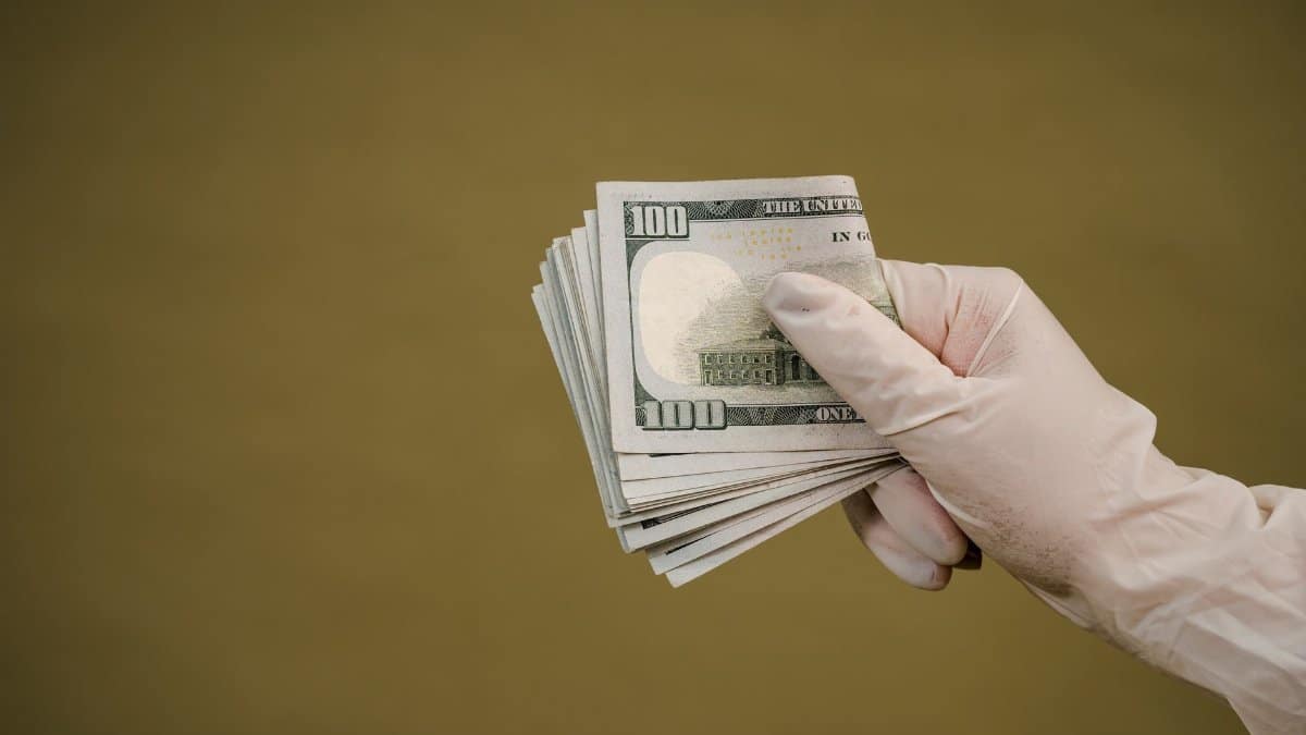 A gloved hand holds a stack of hundred-dollar bills against a plain background.