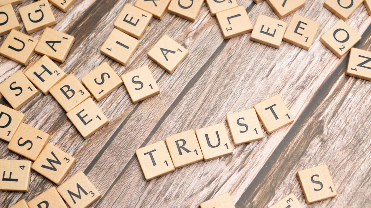 Scrabble letter tiles form the word 'Trust' on a wooden table, surrounded by scattered tiles.