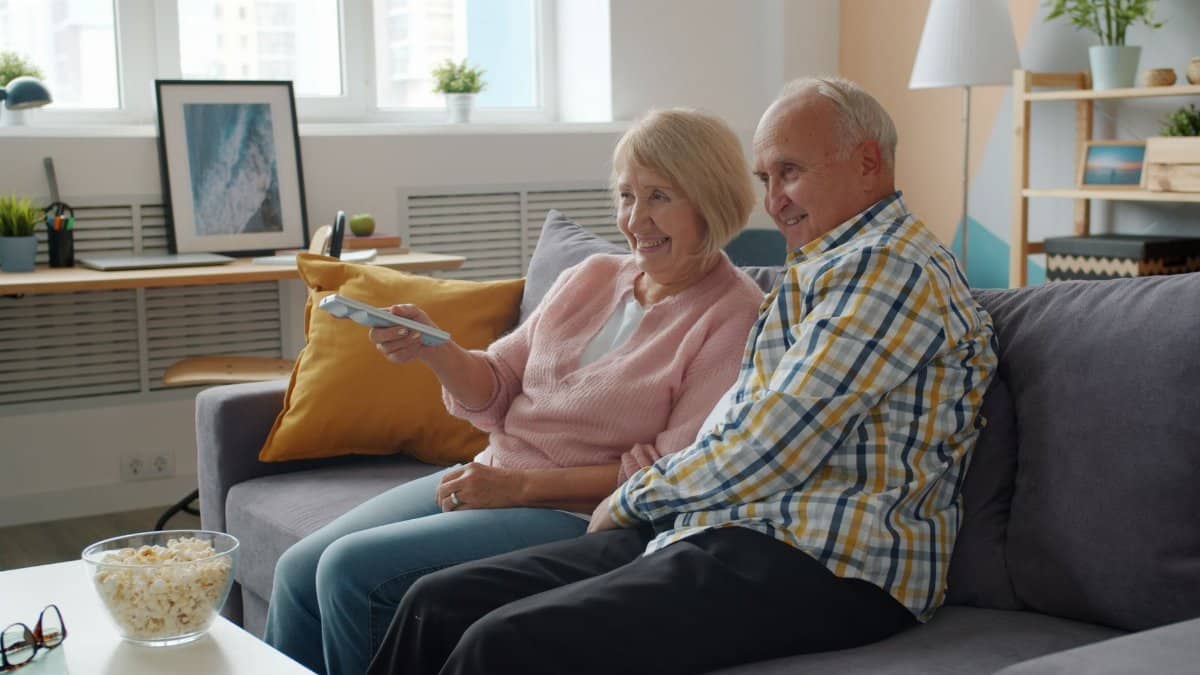 Senior couple watching TV together on a cozy sofa with snacks.