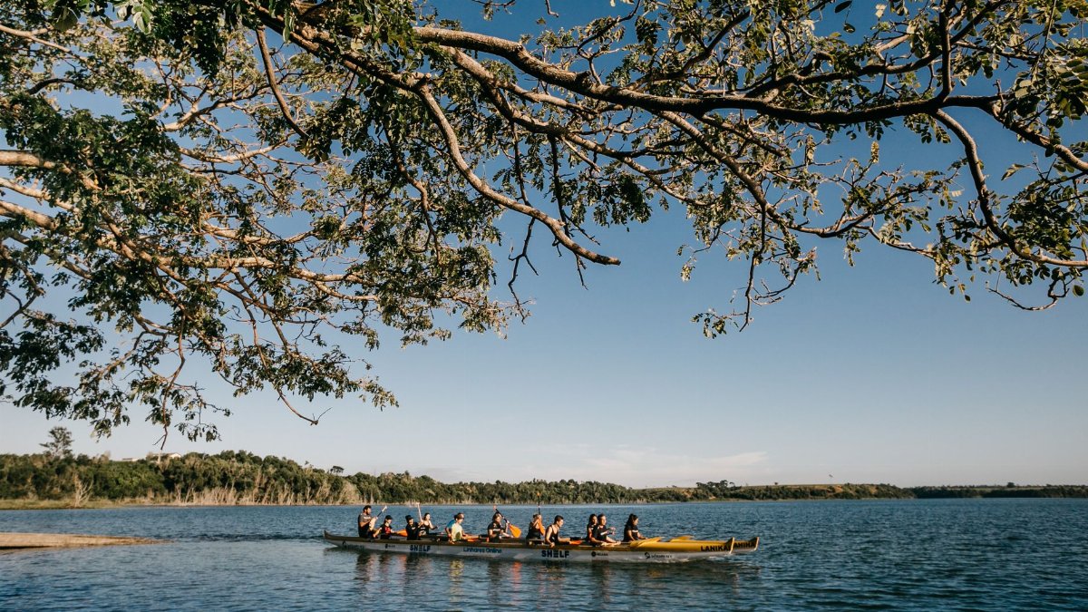 Unrecognizable paddlers in boats reflecting in rippled lake against green trees under cloudy blue sky