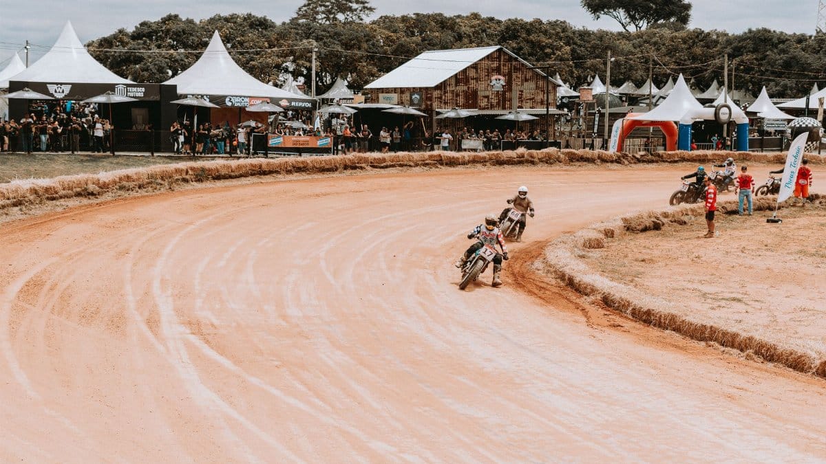 Motorcycle race with riders speeding on a sandy racetrack, surrounded by spectators and tents.