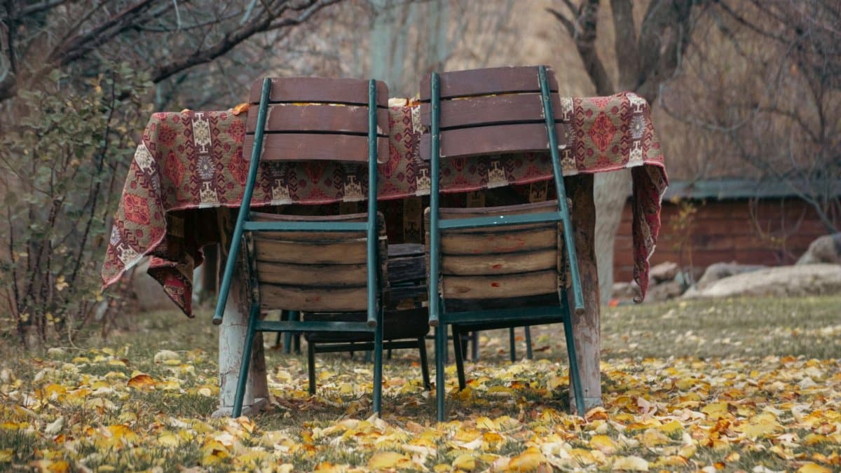 Wooden chairs and table with a patterned cloth surrounded by fall leaves outdoors.