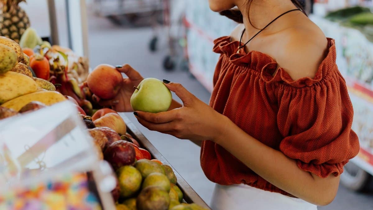 Young woman in orange blouse selecting apples at a bustling outdoor market in daylight.