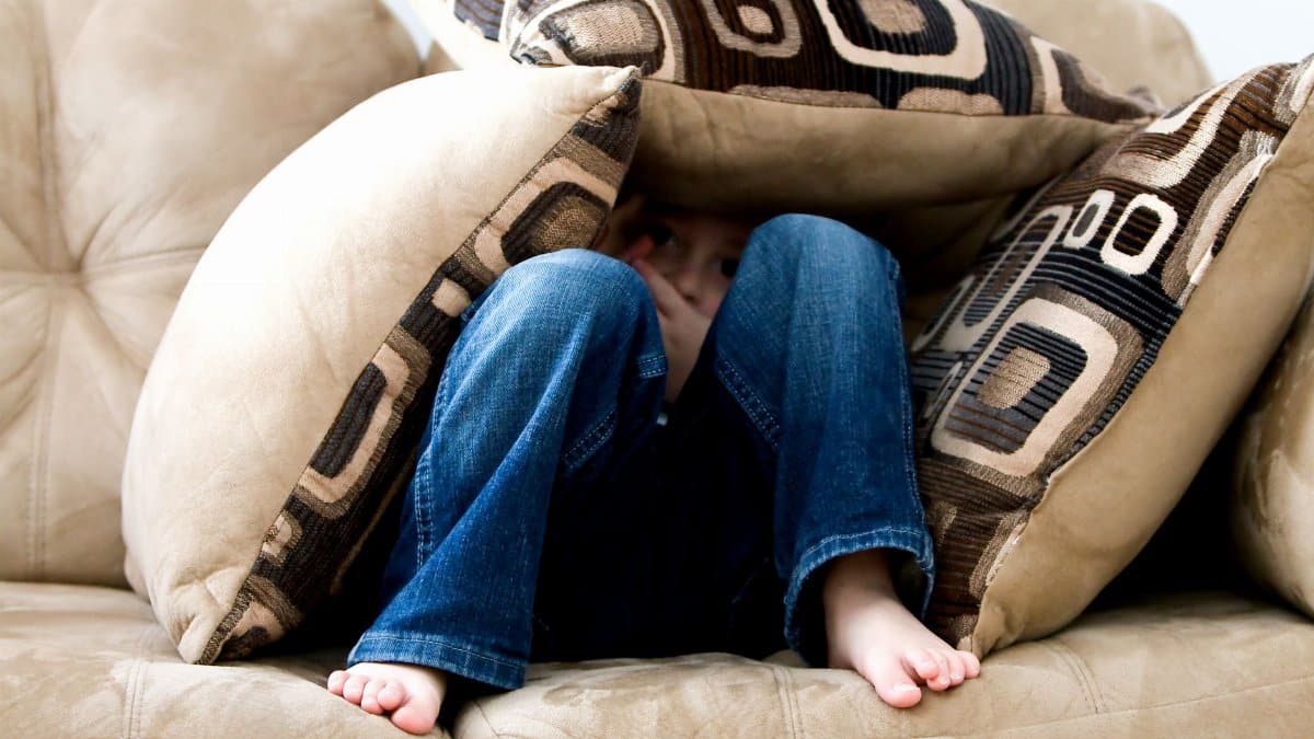 A child crouched under cushions on a couch, conveying a sense of playfulness or fear.