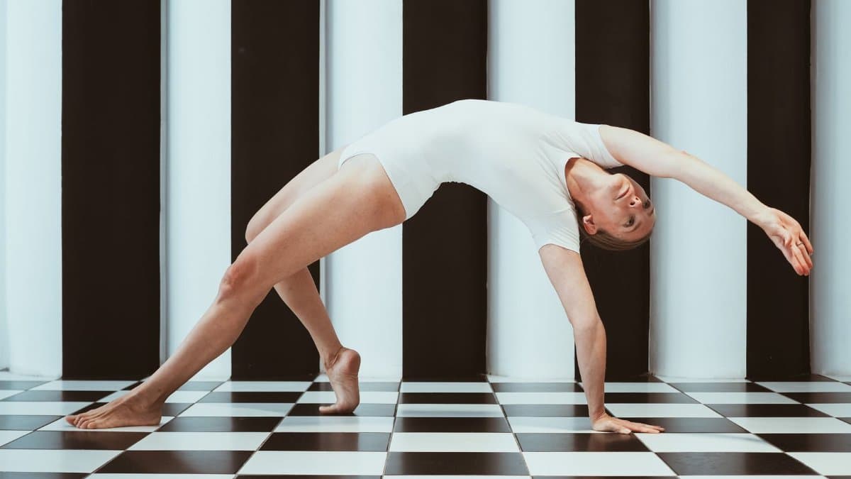 A flexible woman in a white costume performs a gymnastic bridge pose on a checkered floor in a studio.