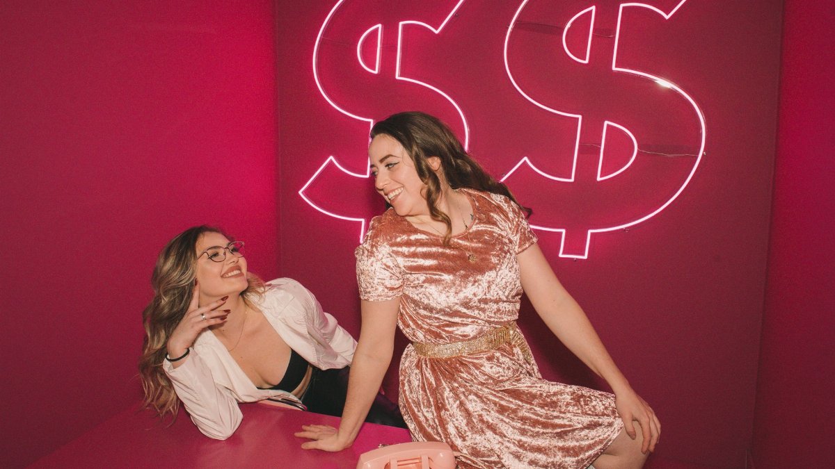Two women enjoying a playful moment in a vibrant pink room with neon dollar signs.
