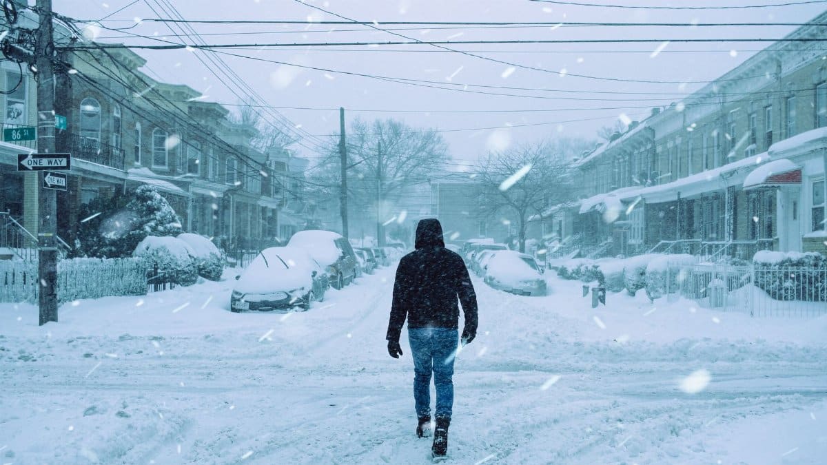 Full body back view of faceless man in outerwear strolling on crossroad on snowy street with parked cars and residential buildings during snowfall