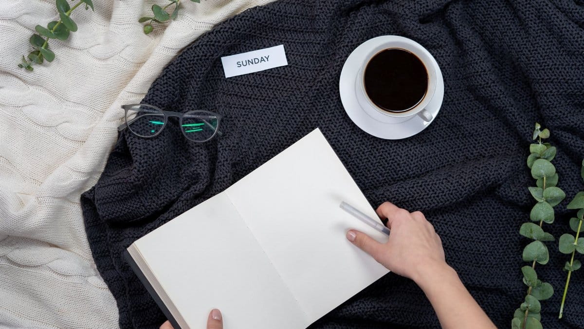 A cozy setup with black coffee, notebook, glasses, and a Sunday note on a knitted blanket.