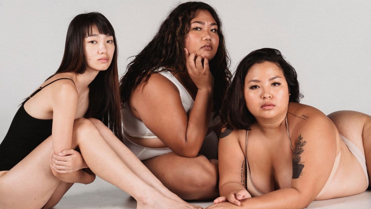 Serious skinny Asian female sitting on floor in studio near plump models in white underwear on white background and looking at camera
