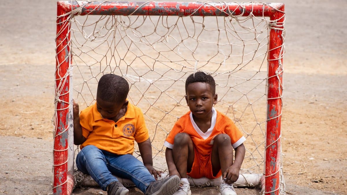 Two young African boys sitting inside a small soccer goal outdoors, wearing orange shirts.