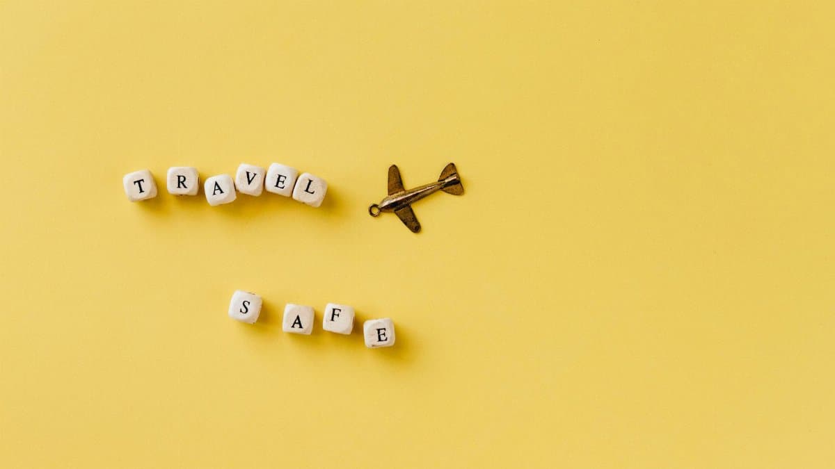 Flat lay of 'Travel Safe' text with airplane pendant on yellow background, evoking travel and safety concepts.