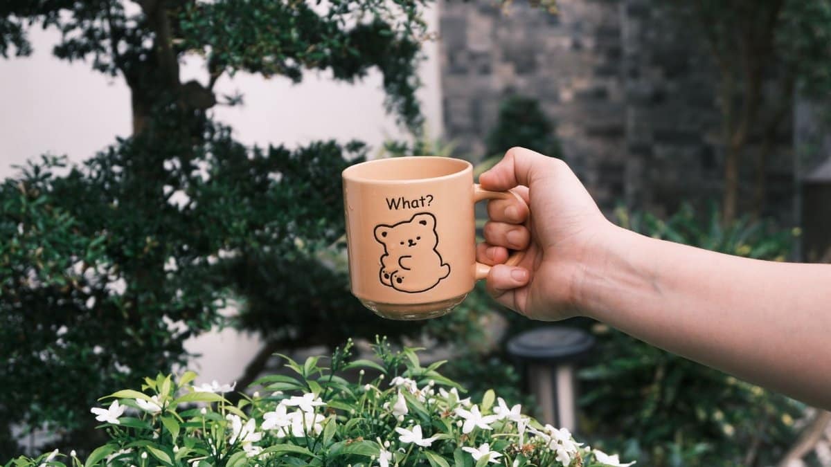 Hand holding a bear-themed mug in a garden setting with lush greenery.