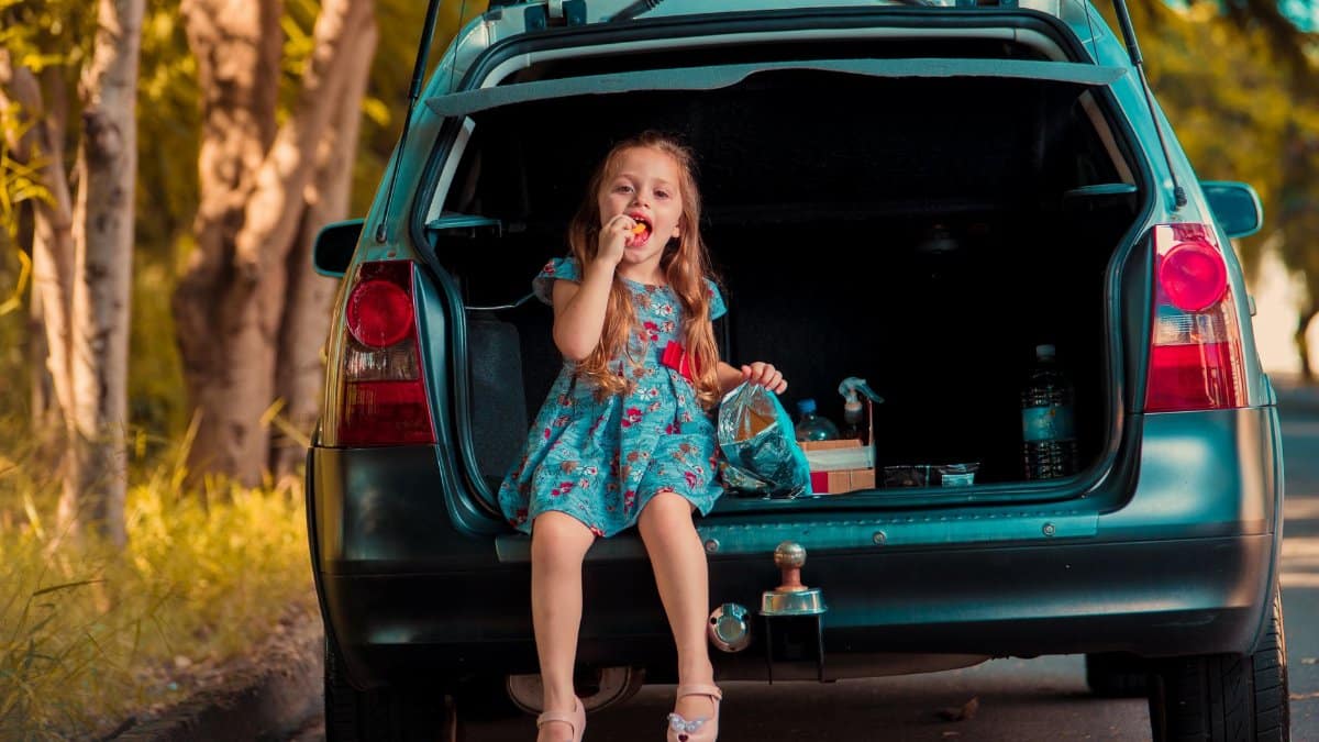 A young girl in a floral dress enjoys a snack sitting in the trunk of a car on a sunny day.