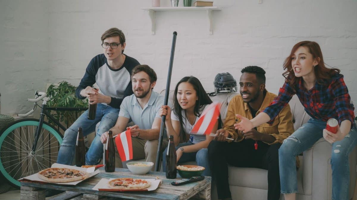 Group of diverse friends cheering for a game at home with snacks and flags.