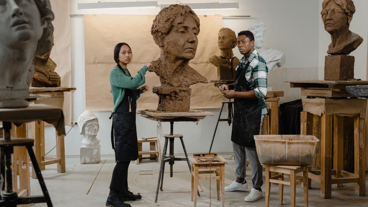 Two artists work on clay sculptures in a well-lit art studio.
