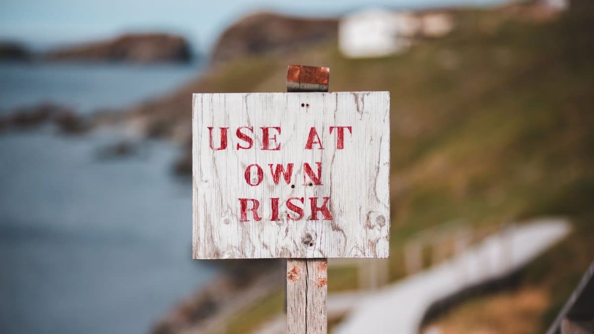 Weathered poled sign with inscription Use At Own Risk located on stiff cliff slope near staircase against blurred sea