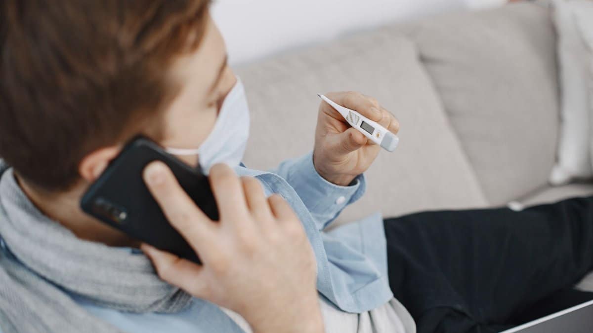 A man checks his temperature while on a telehealth call from home, wearing a face mask for safety.