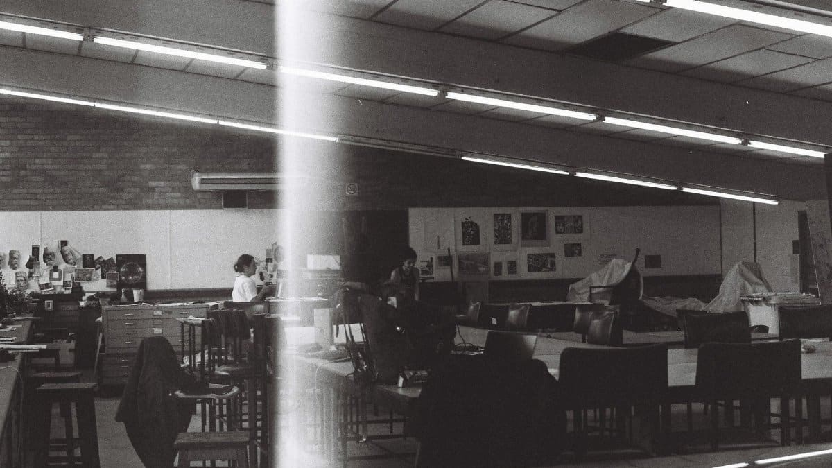 Moody black and white image showcasing an empty art classroom with people at desks.