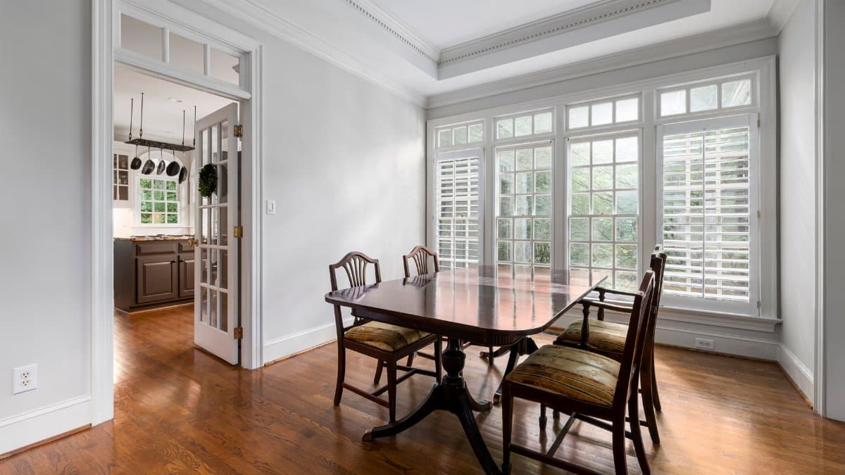 Bright dining room featuring a wooden table, classic chairs, and large windows overlooking greenery.