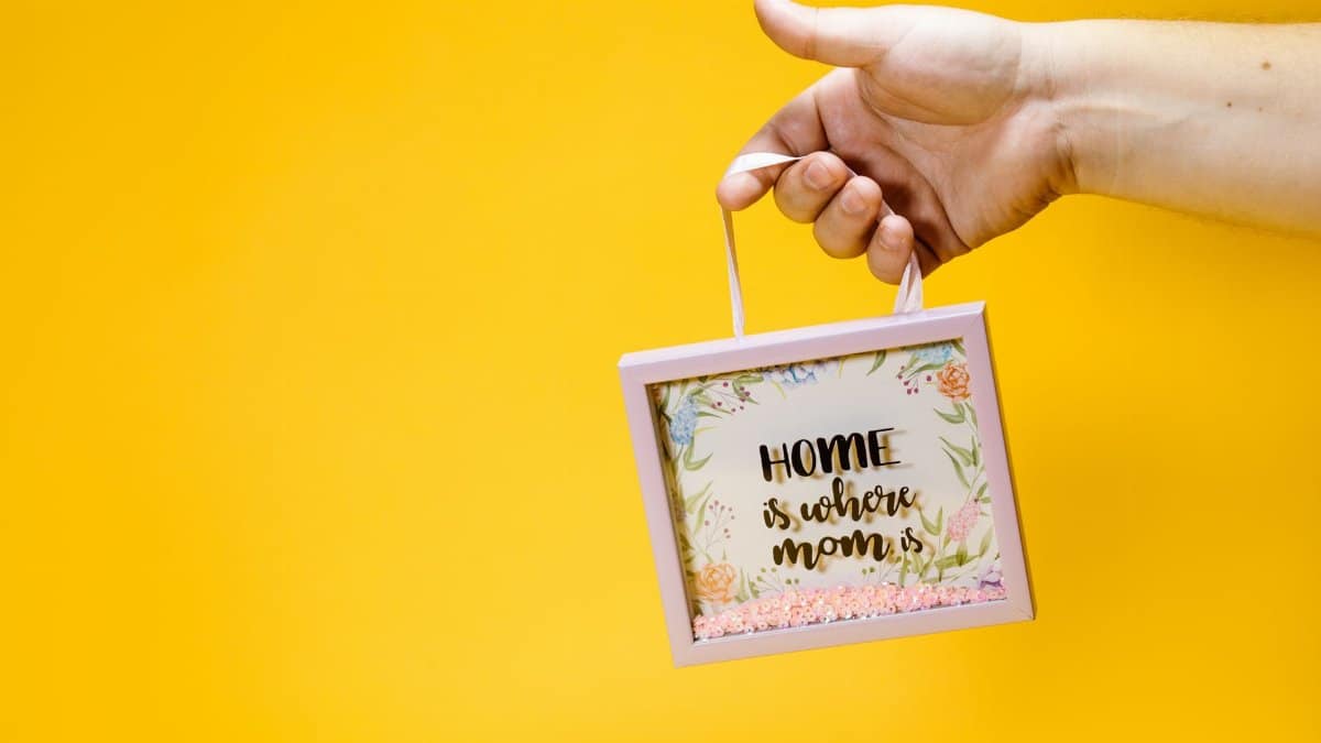 A hand holding a floral picture frame with 'Home is where mom is' text against a vibrant yellow background.