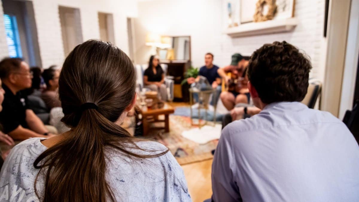 A warm and casual indoor gathering features people socializing, with music being played in a living room setting.