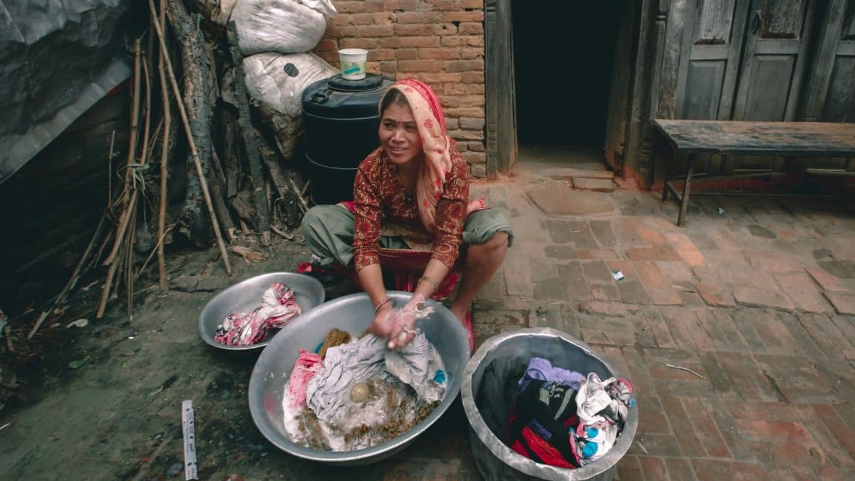 From above of content young ethnic female in traditional wear washing clothes in metal basins while looking away against old house