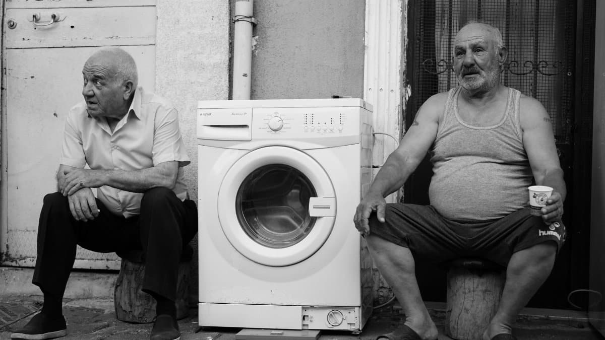 Two men seated outside, casually sitting with a washing machine between them.