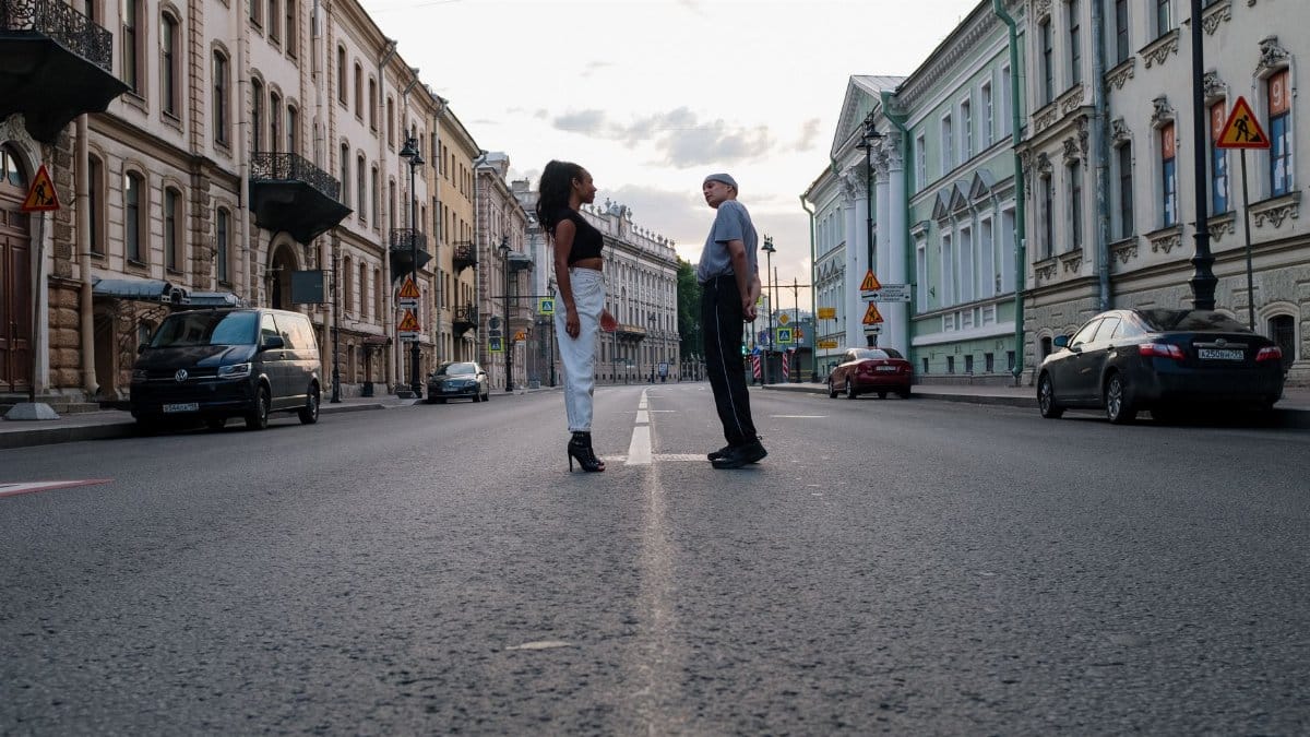 A pair of people face each other on an empty urban street lined with classic architecture.