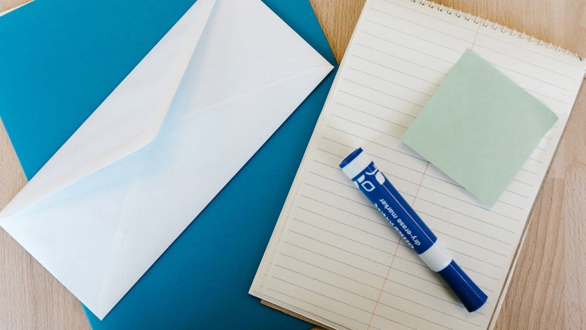 Flat lay of office supplies on a wooden desk, including a notepad, envelope, marker, and sticky note.