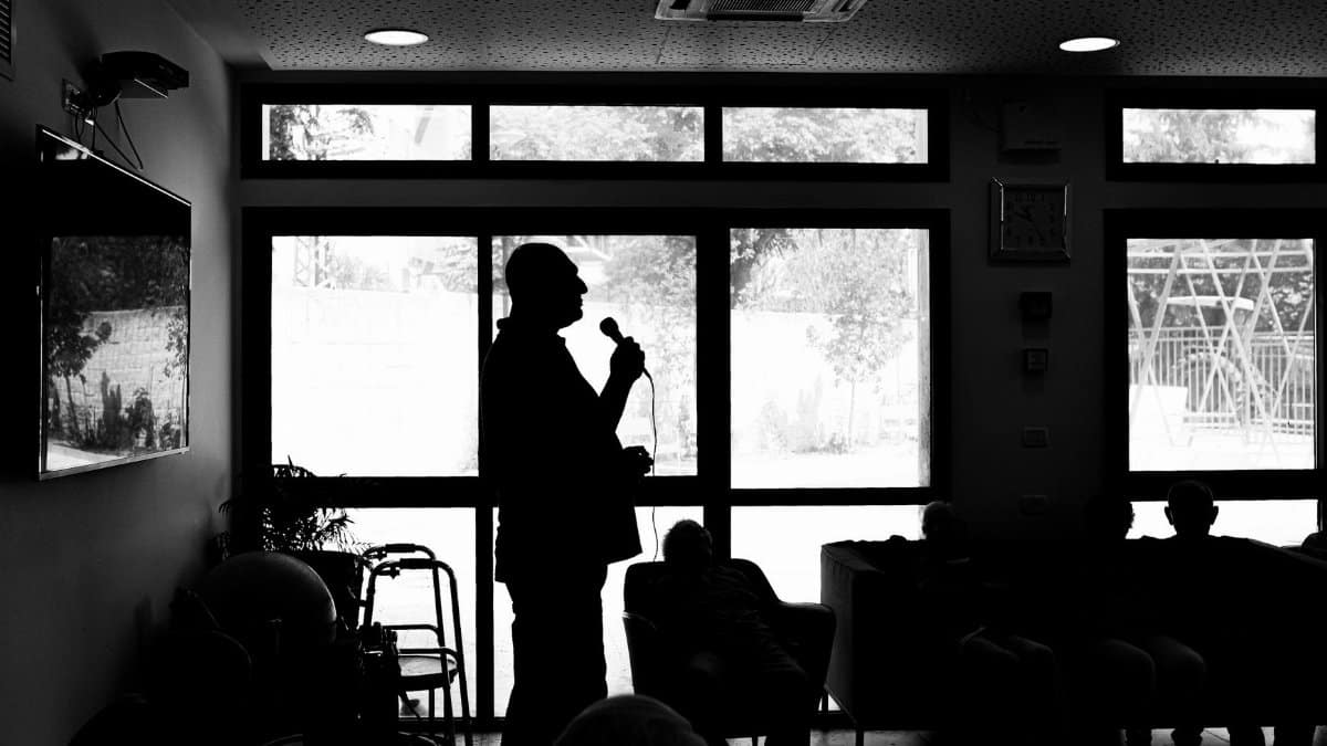 Silhouette of a person speaking indoors, with an attentive audience in the shadows.