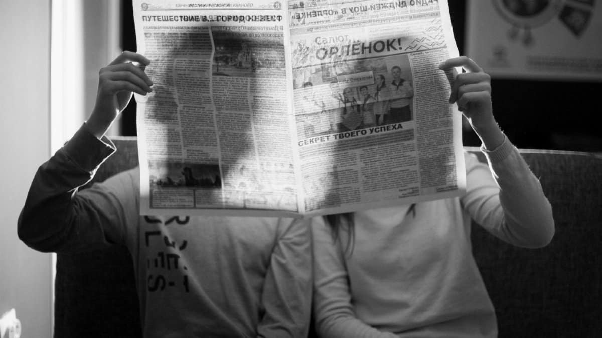 A couple sitting indoors reading a newspaper in a black and white photograph.