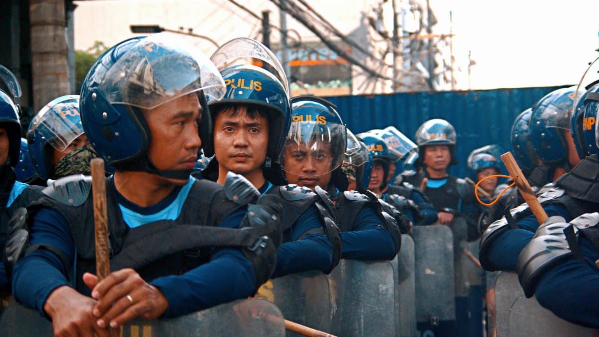 Riot police officers lined up with helmets and shields, maintaining order in a city setting.