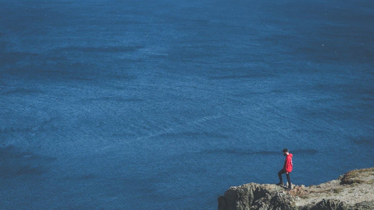 A man in a red jacket stands on a cliff edge overlooking a vast ocean.