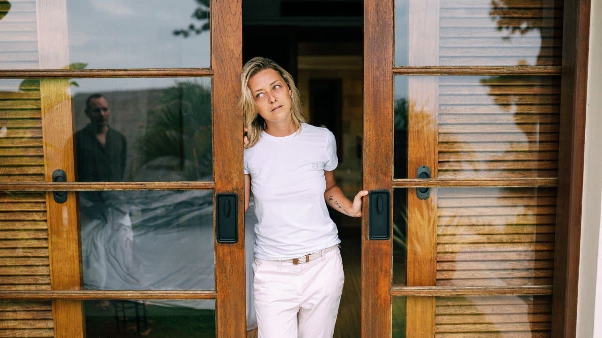 A woman in a white shirt stands by a wooden sliding door, with a reflection visible.