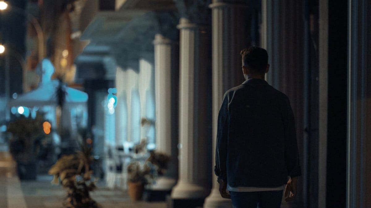 A person walks at night along a city sidewalk with pillars, creating a moody atmosphere.
