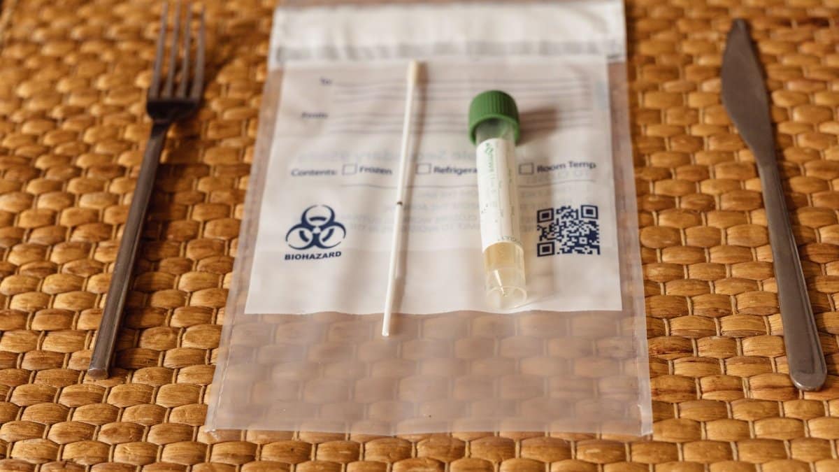 A Covid-19 testing kit with swab and test tube on a straw placemat between fork and knife.