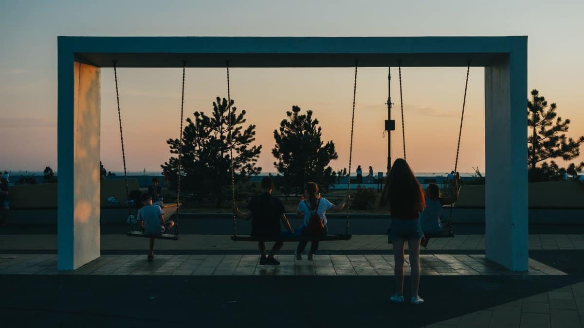 A group of teenagers relaxing on swings at dusk near the coastline, silhouetted against a colorful sunset sky.