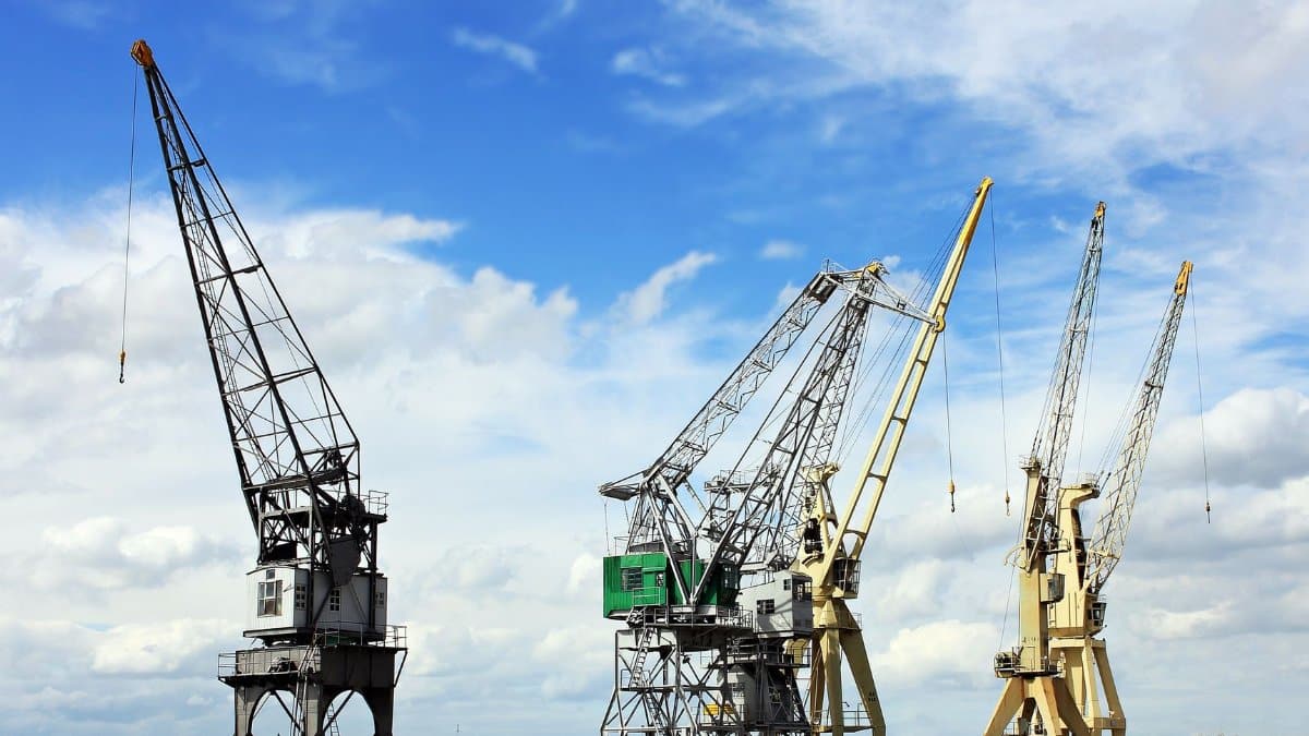 Industrial cranes reaching into a bright blue sky with scattered clouds, a symbol of construction and industry.